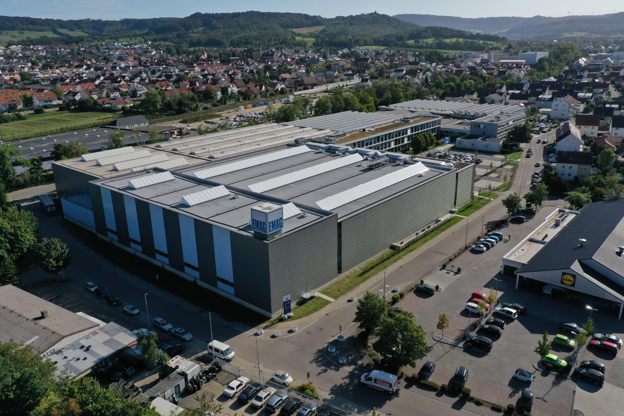 Die dunkle Montagehalle mit dem EMAG-Schriftzug auf dem Dach, dahinter der Ortskern von Salach