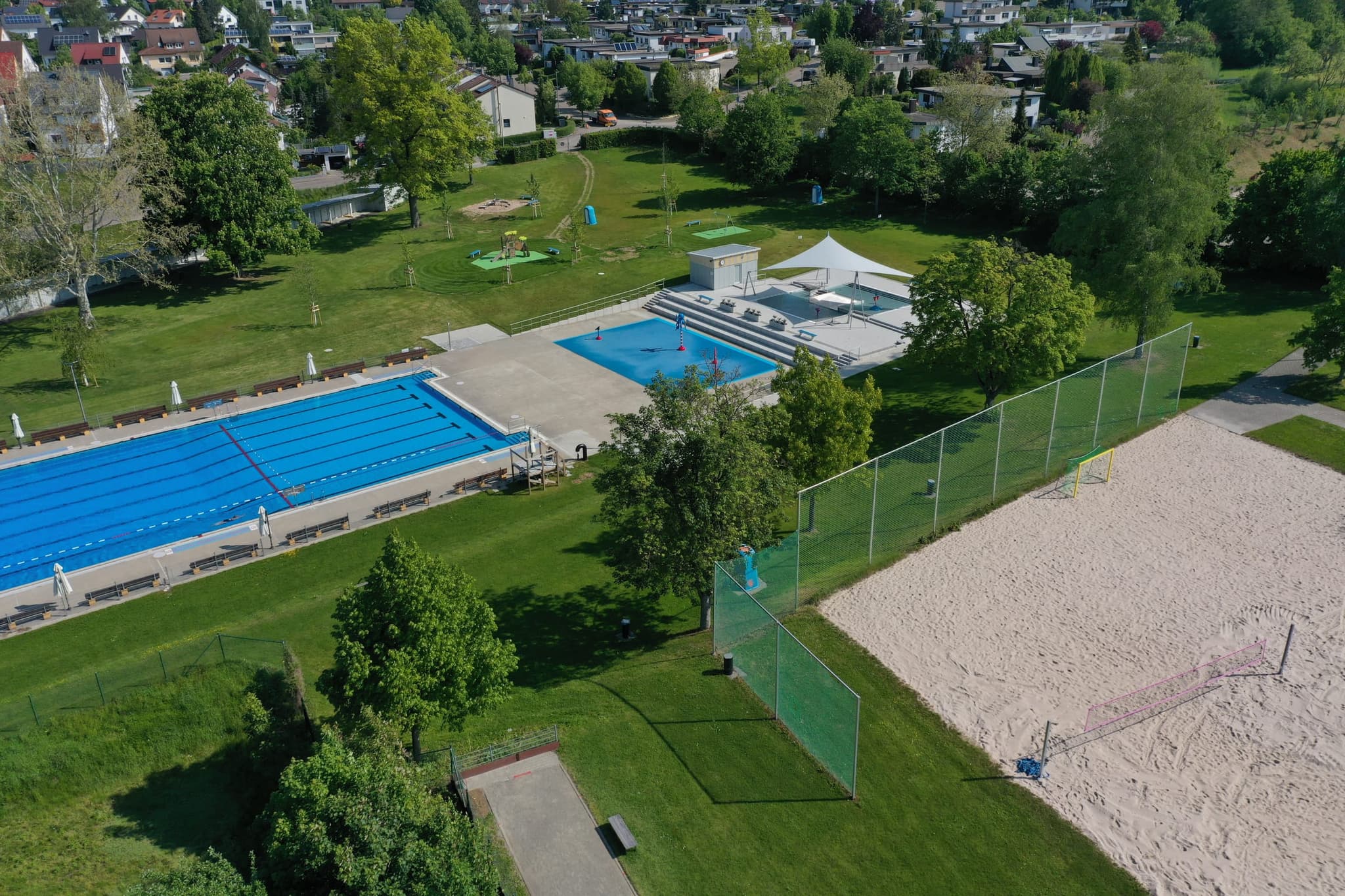 Beide Becken und das Beachvolleyballfeld nebeneinander, dahinter die Wohnbebauung